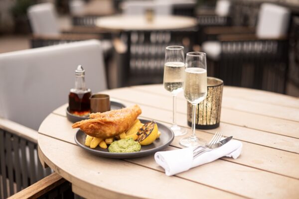 Two champagne flutes on a wooden outdoor table beside a plate of fried food and sides, with a blurred restaurant patio in the background