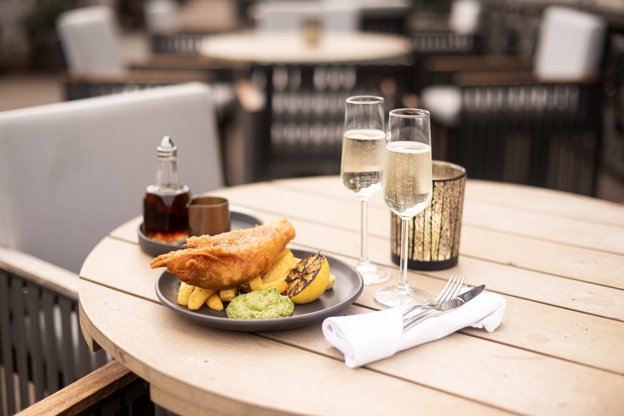 Two champagne flutes on a wooden outdoor table beside a plate of fried food and sides, with a blurred restaurant patio in the background