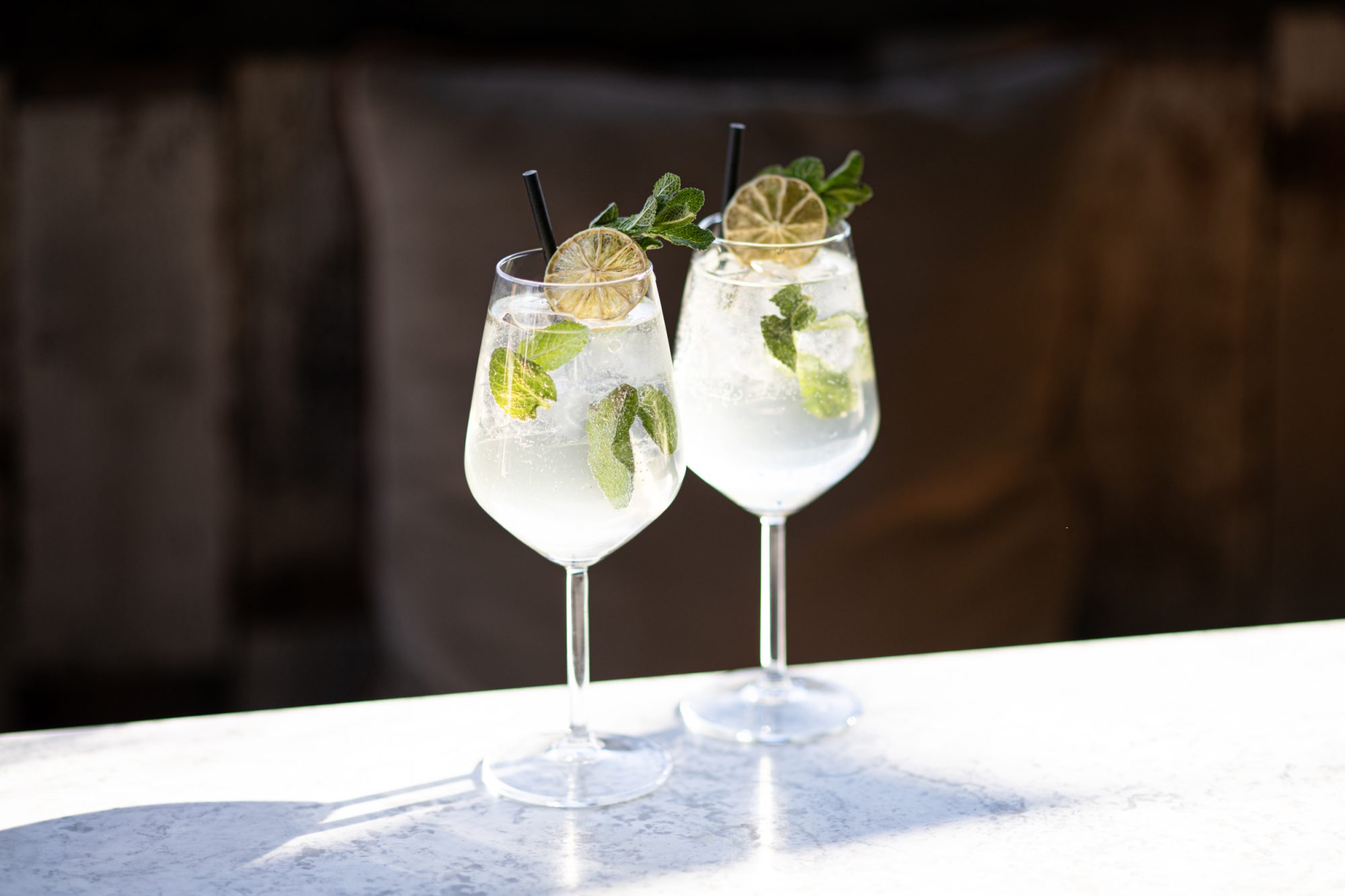Two stemmed cocktails on a white marble bar, garnished with lime wheels, mint, and black straws.