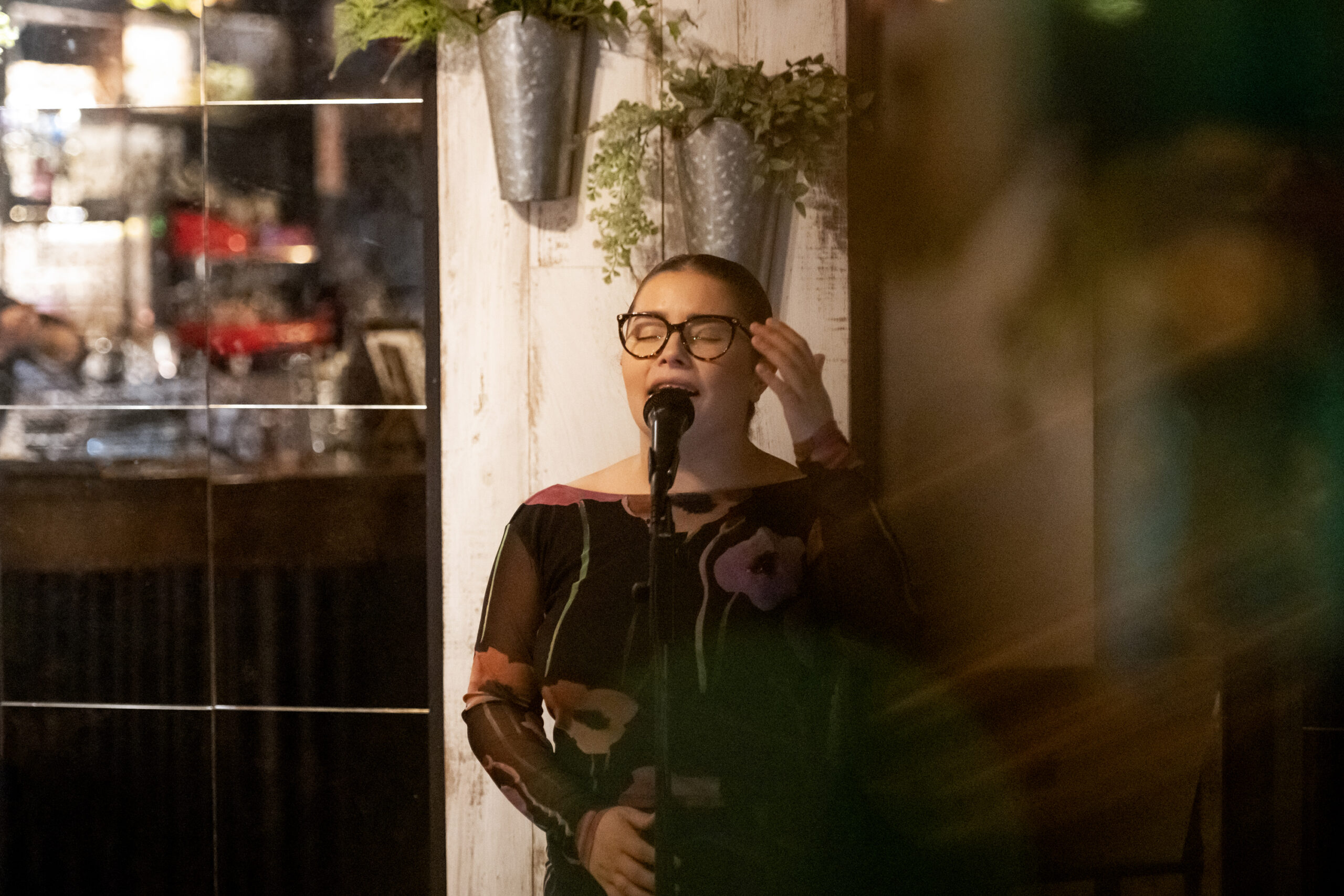 Person with short hair and glasses singing into a microphone on a stage with a wooden wall and hanging plants behind them.