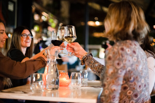 Friends toast with wine glasses at a candlelit restaurant table, clinking glasses in a warm, social moment.