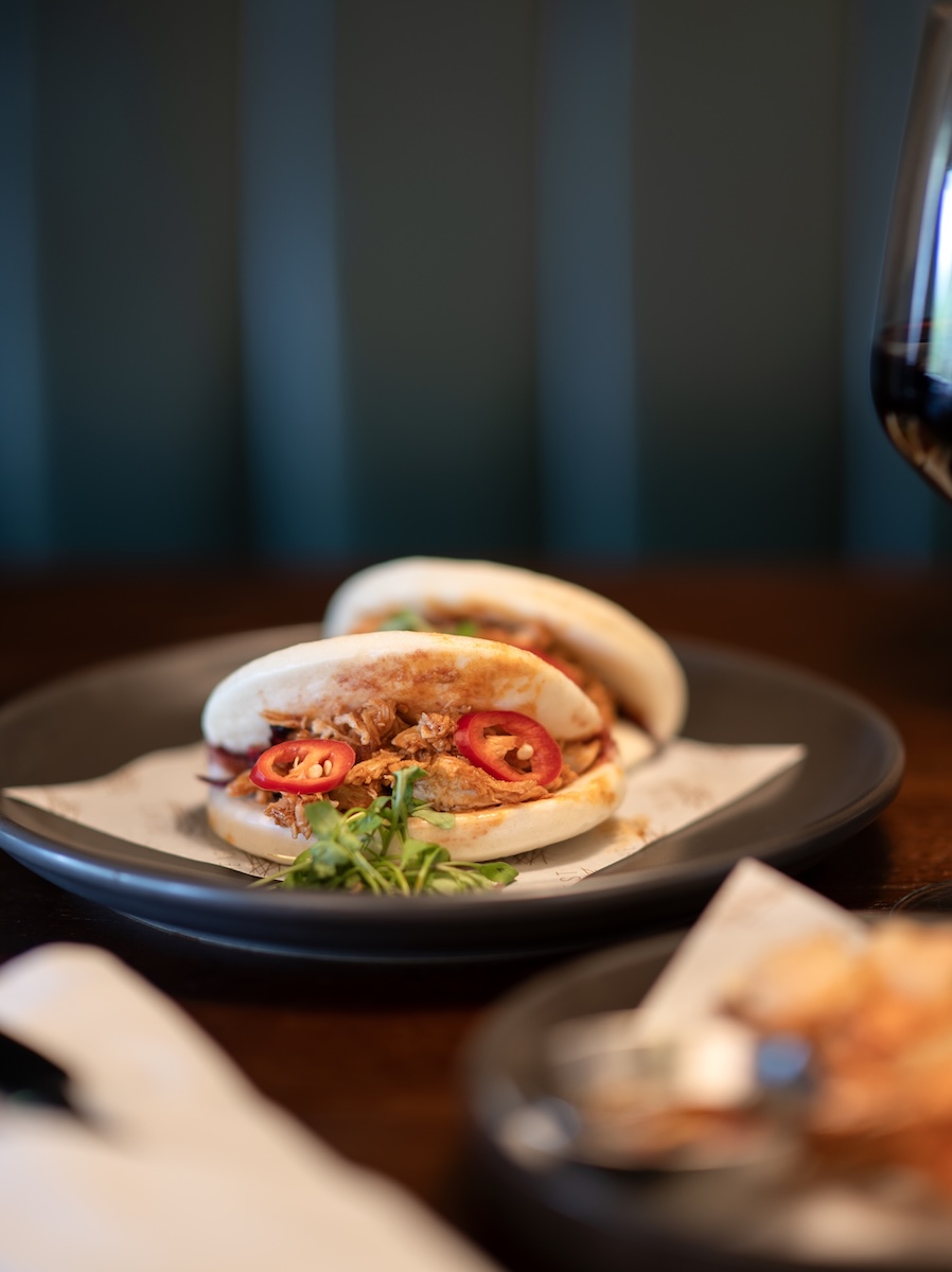 Two soft tacos filled with shredded meat, sliced red chili, and cilantro on a dark plate with a glass of red wine nearby.
