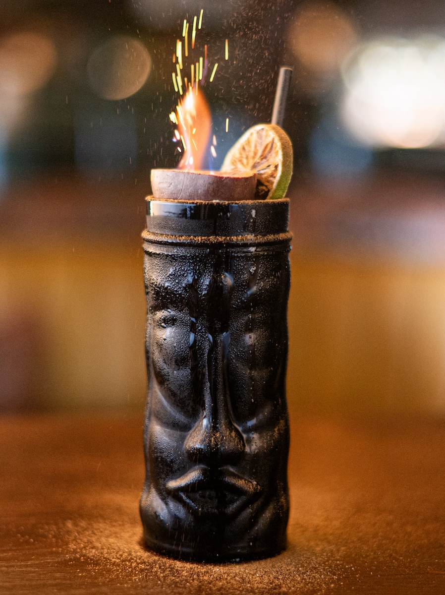 Black tiki mug holds a flaming cocktail with a lime wedge and a straw on a bar surface.