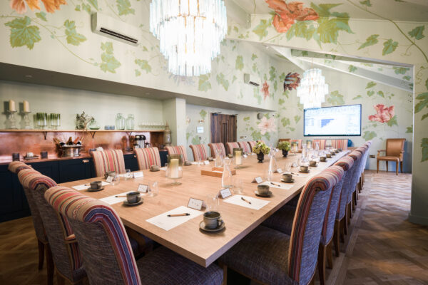 Large conference room with a long wooden table set for a meeting, striped chairs, floral wallpaper, and a chandelier.