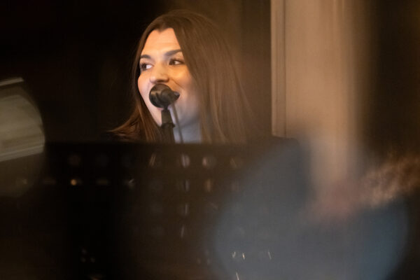 Woman singing into a microphone on stage, with a blurred foreground and warm lighting.