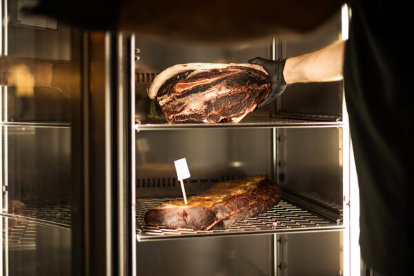 Hand wearing black glove places a large slab of raw beef into a refrigerated display case in a butcher shop setting.