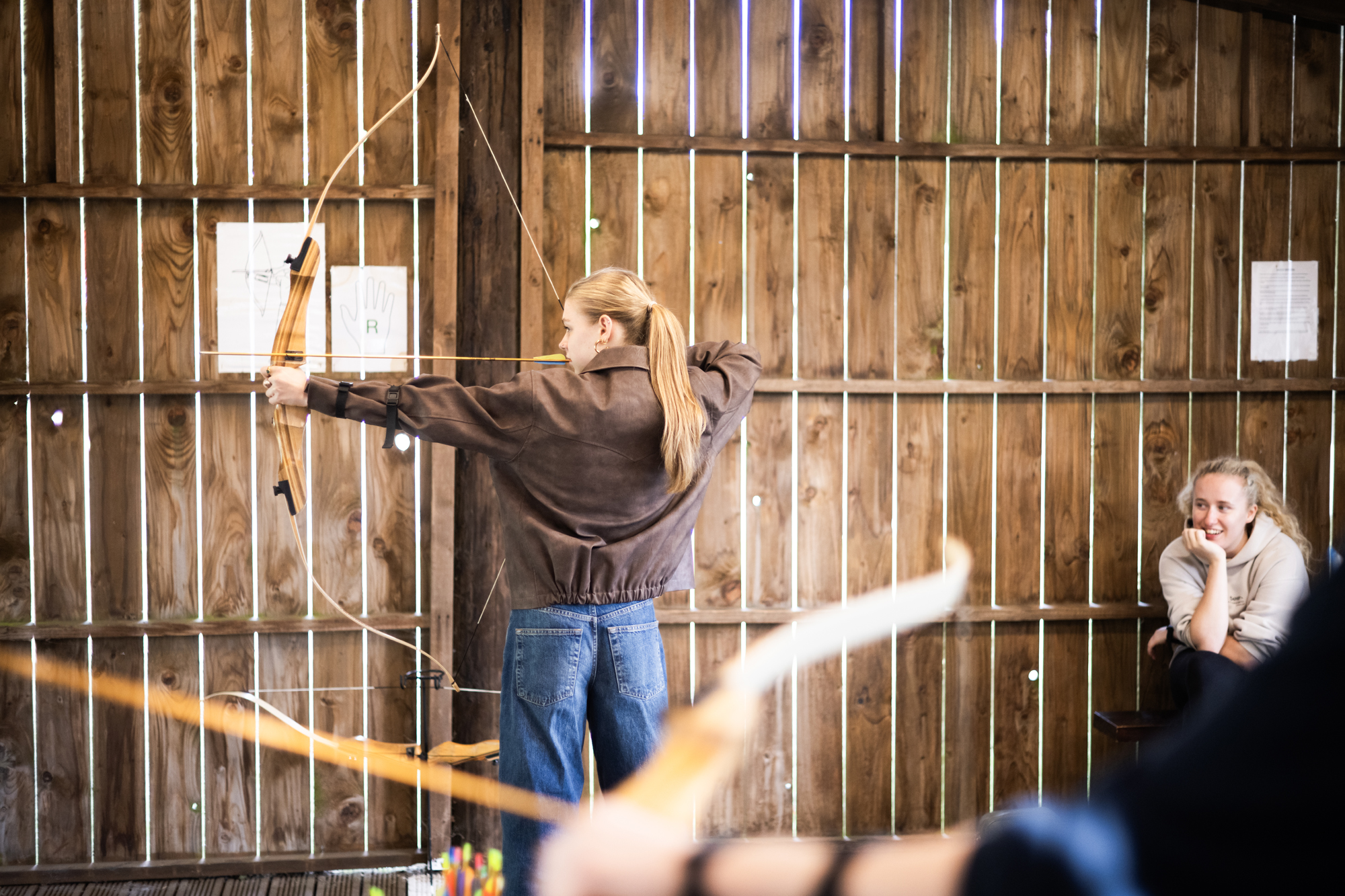 Young woman draws a bow indoors in a wooden shooting space, aiming at a target while others watch.