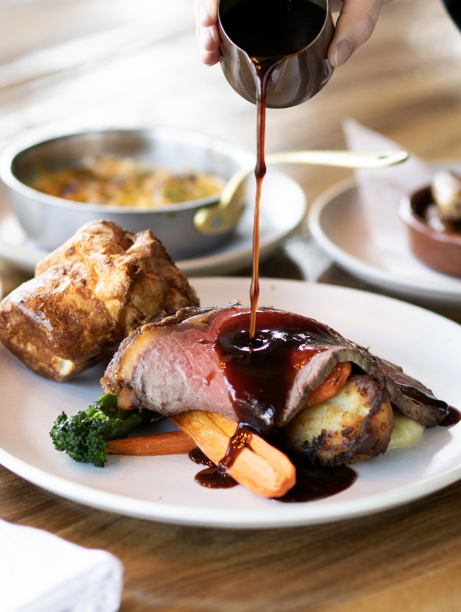 Sliced medium-rare beef tenderloin being coated with a dark glaze atop roasted vegetables on a white plate, with crusty bread nearby.