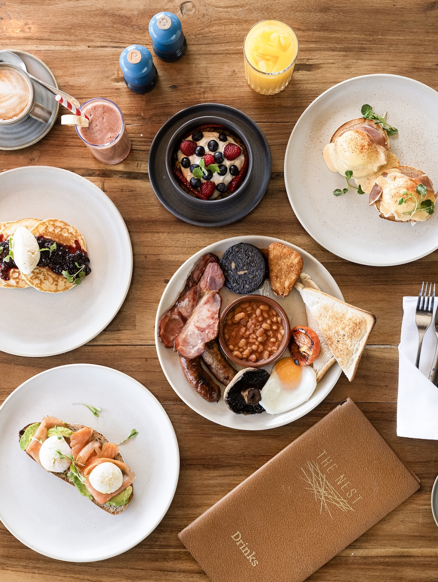 Overhead view of a brunch spread on a wooden table: central beans and toast plate surrounded by pancakes, smoked-salmon toast, croissants, berry yogurt, and a plate of berries with pastry and drinks nearby