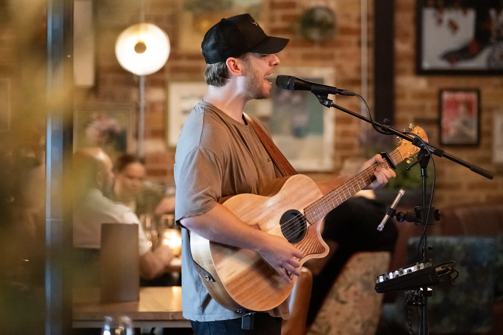 Male musician in a cap sings into a microphone while playing an acoustic guitar in a cozy cafe setting.