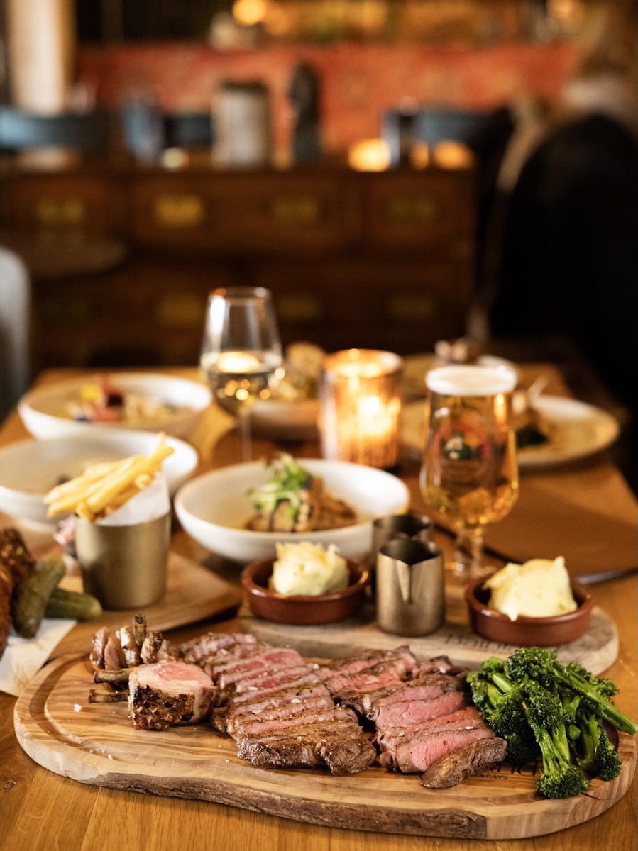 Sliced medium-rare steak on a wooden board with broccoli, mashed potatoes, and sauces at a dimly lit restaurant table.