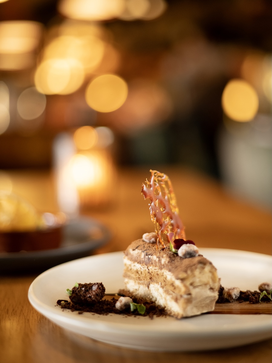 Dessert slice on a white plate with chocolate crumble and a delicate caramel lattice, warm bokeh lights in the background.