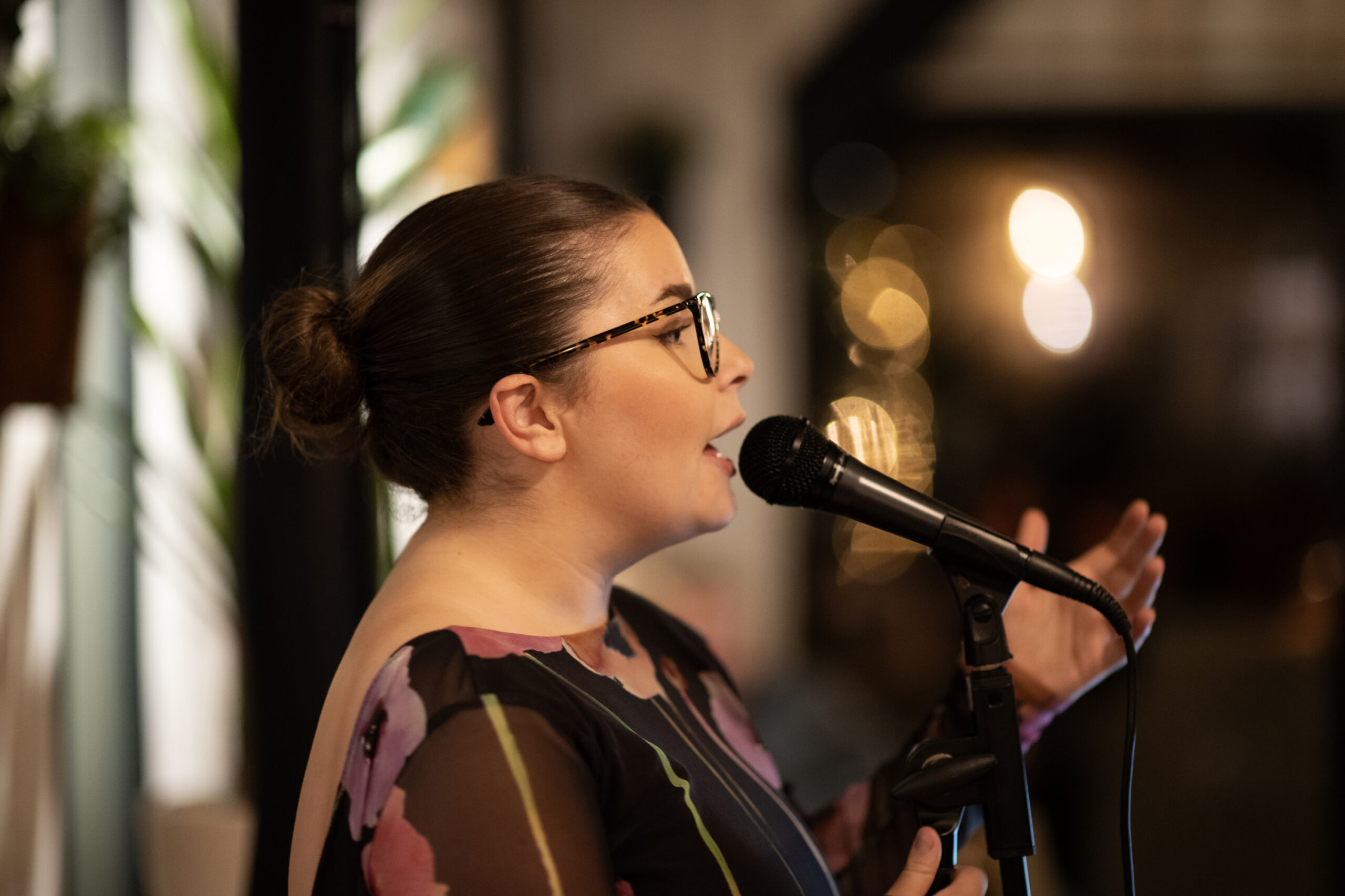 Side profile of a woman singing into a microphone, wearing tortoiseshell glasses and a patterned dress or top.